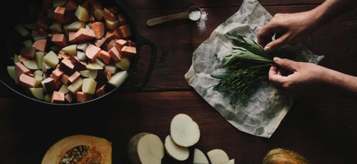 Coup de frais généraux d'une table en bois avec un grand pot en fonte avec des légumes-racines coupés, une demi-courge et des pommes de terre en tranches. Il y a un emballage ouvert d'emballage alimentaire en cire d'Abeego contenant des herbes